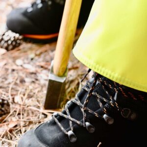 SwedePro Chainsaw Protector Boot being worn, first third of boots shown the rest covered by yellow chainsaw protector chaps with an axe propped between the feet