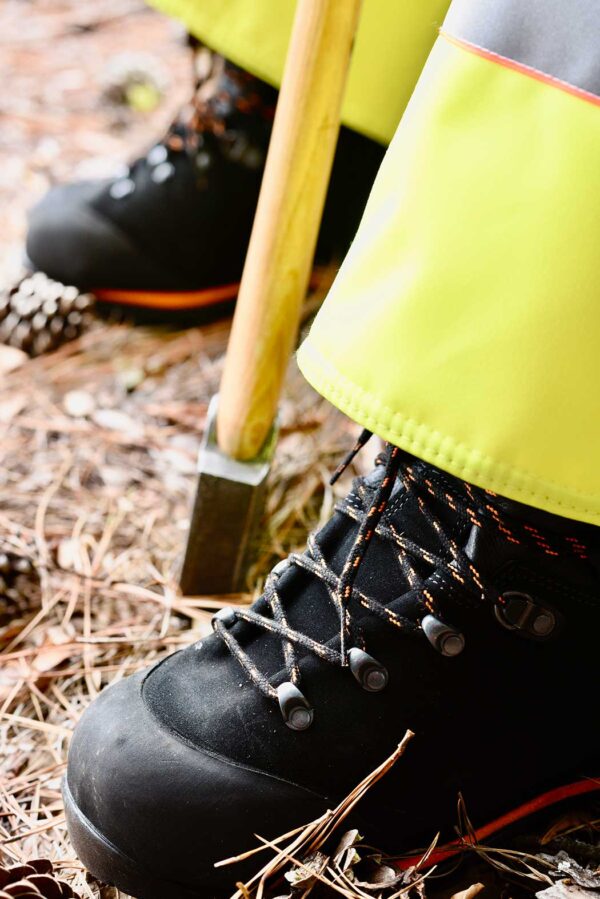 SwedePro Chainsaw Protector Boot being worn, first third of boots shown the rest covered by yellow chainsaw protector chaps with an axe propped between the feet